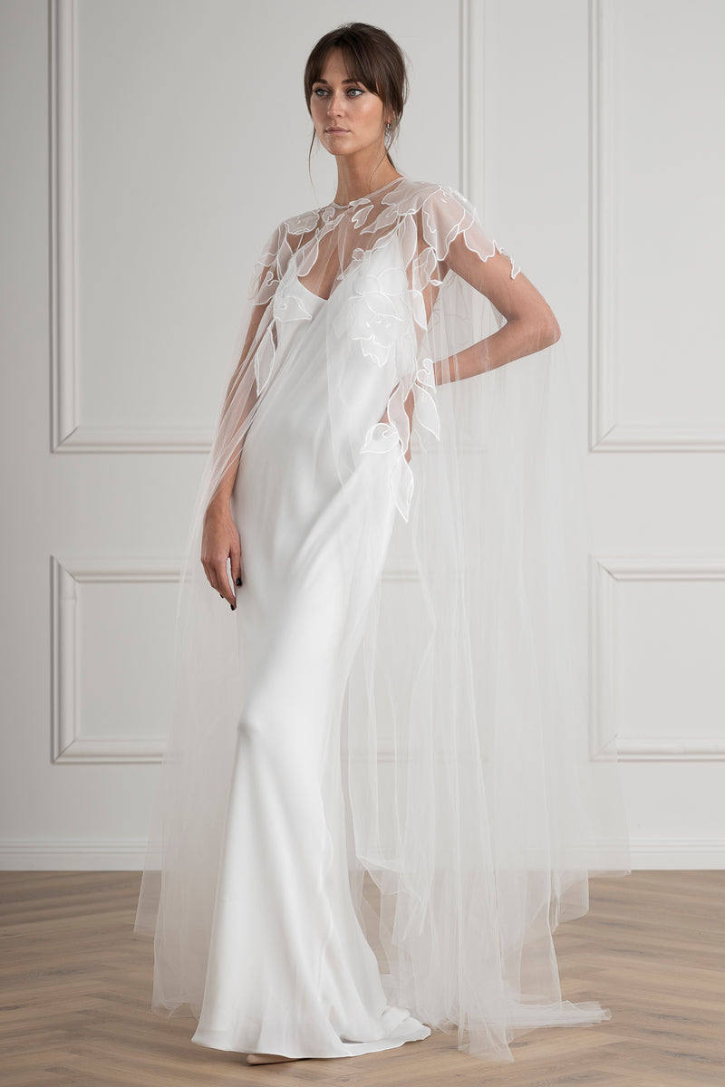 Bride wearing a white slip  wedding dress with sheer sleeves against a white paneled wall.